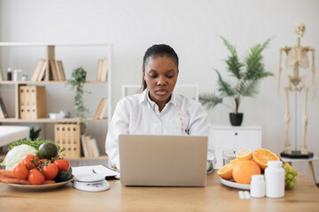 Canvas Print - Attractive multiracial female in white coat working on portable computer in cozy workplace at modern hospital. Mindful dietitian reading about latest researches in nutrition science via internet.