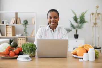 Canvas Print - Attractive multiracial female in white coat working on portable computer in cozy workplace at modern hospital. Mindful dietitian reading about latest researches in nutrition science via internet.