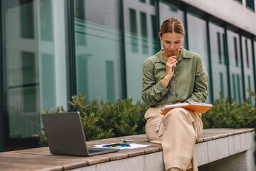 Wall Mural - Pensive woman entrepreneur working on laptop outside on modern building background and making notes