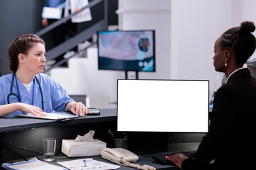 Wall Mural - African american receptionist using pc with white isolated screen at reception counter desk in hospital. Diverse medical staff working with mockup template during checkup visit consultation