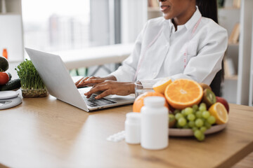 Canvas Print - Focus on hands multicultural lady in doctor's coat typing on modern laptop in consulting room of medical center. Experienced nutrition professional searching for weight loss information.