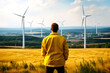© Garnar - Back view of a male engineer in a yellow work jacket looking out at a field with a wind turbine farm in front of him. The concept of people in a community with sustainable resources. Renewable energy