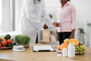 Wall Mural - Focus on office desk with paper clipboard near products and medicine on background of lady's visit to nutritionist. African american expert and client promoting wholesome lifestyle via healthcare.