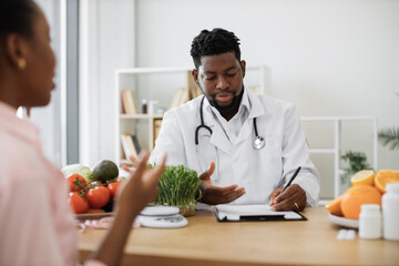 Wall Mural - Confident man with stethoscope writing on paper clipboard during conversation with multiracial woman in office. Serious nutrition expert making notes of patient's medical history for proper meal plan.