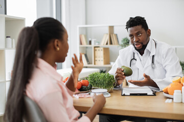 Wall Mural - Focused african american man in white coat holding avocado while talking with female client in consulting room. Medical specialist in food encouraging to enriching eating with fruit full of nutrients.