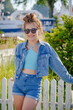 Vertical portrait of stylish smiling little girl in sunglasses stands against backdrop of nature and home. Girl is dressed in blue denim clothes