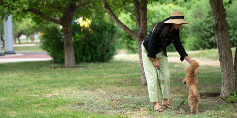 An Asian girl (Kazakh woman) feeds her dog (mini poodle) from her hands. Summer portrait of a young woman in the park.