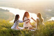 Three beautiful young caucasian ladies with wine in their hands enjoy spending time on picnic at summer sunset. Good weekend concept with friends	