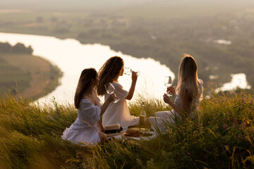 Wall Mural - Girlfriends having picnic on the mountain at sunset. Group of young women sitting on grass on summer evening. Girlfriends drinking wine on outdoor party.	
