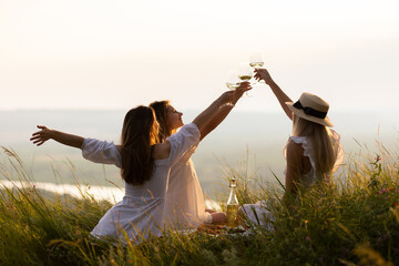 Wall Mural - Three young beautiful smiling female in summer white dresses. Carefree women making picnic in the park at sunset. Positive models sitting on plaid on grass, drinking wine, eating fruits and cheese.	
