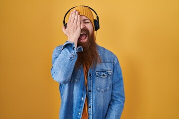 Canvas Print - Caucasian man with long beard listening to music using headphones covering one eye with hand, confident smile on face and surprise emotion.