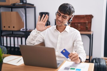 Wall Mural - Young hispanic man working using computer laptop holding credit card waiving saying hello happy and smiling, friendly welcome gesture