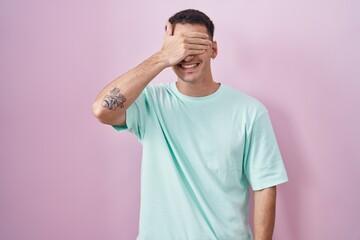 Wall Mural - Handsome hispanic man standing over pink background smiling and laughing with hand on face covering eyes for surprise. blind concept.