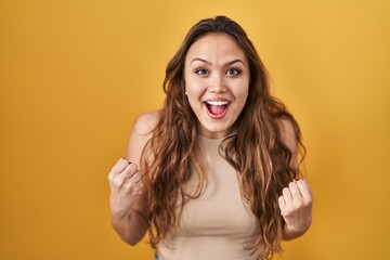 Sticker - Young hispanic woman standing over yellow background celebrating surprised and amazed for success with arms raised and open eyes. winner concept.