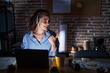 Wall Mural - Beautiful brunette woman working at the office at night smiling with happy face looking and pointing to the side with thumb up.