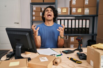 Poster - Hispanic man with curly hair working at small business ecommerce crazy and mad shouting and yelling with aggressive expression and arms raised. frustration concept.