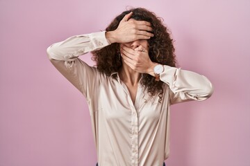 Sticker - Hispanic woman with curly hair standing over pink background covering eyes and mouth with hands, surprised and shocked. hiding emotion