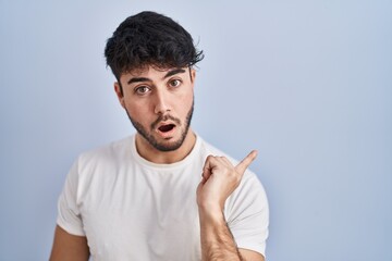 Wall Mural - Hispanic man with beard standing over white background surprised pointing with finger to the side, open mouth amazed expression.