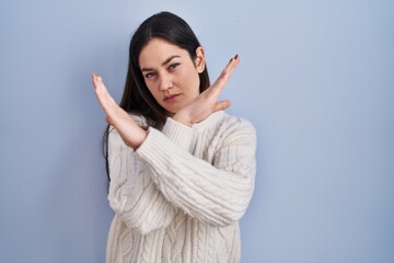 Sticker - Young brunette woman standing over blue background rejection expression crossing arms doing negative sign, angry face