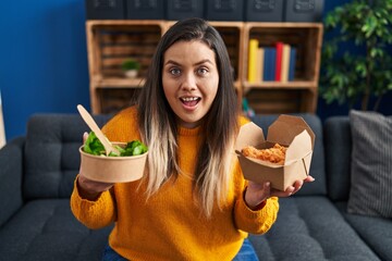 Poster - Young hispanic woman holding healthy salad and fried chicken wings celebrating crazy and amazed for success with open eyes screaming excited.