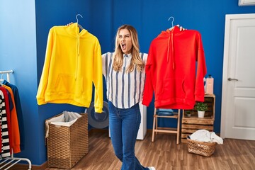 Sticker - Young blonde woman holding clean laundry on hangers angry and mad screaming frustrated and furious, shouting with anger. rage and aggressive concept.