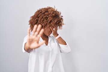 Sticker - Young hispanic woman with curly hair standing over white background covering eyes with hands and doing stop gesture with sad and fear expression. embarrassed and negative concept.