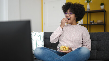 Poster - Young african american woman watching tv sitting on sofa eating chips at home