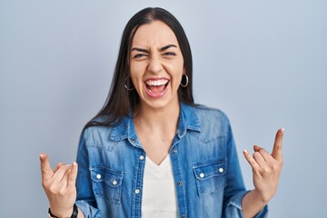 Canvas Print - Hispanic woman standing over blue background shouting with crazy expression doing rock symbol with hands up. music star. heavy music concept.