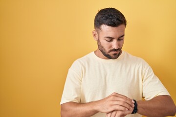 Sticker - Handsome hispanic man standing over yellow background checking the time on wrist watch, relaxed and confident