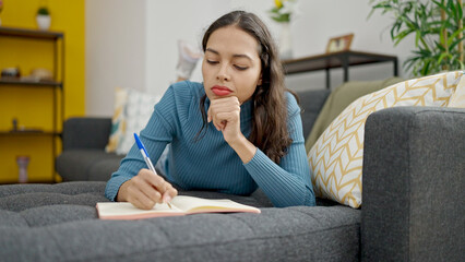 Poster - Young beautiful hispanic woman writing on notebook lying on sofa at home