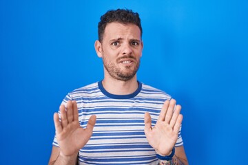 Poster - Young hispanic man standing over blue background moving away hands palms showing refusal and denial with afraid and disgusting expression. stop and forbidden.