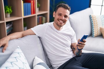 Poster - Young hispanic man using smartphone sitting on sofa at home