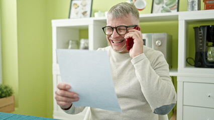 Canvas Print - Middle age grey-haired man talking on smartphone reading document at home