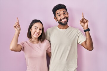 Poster - Young hispanic couple together over pink background smiling amazed and surprised and pointing up with fingers and raised arms.