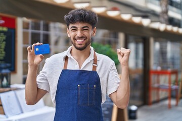 Sticker - Arab man with beard wearing waiter apron at restaurant terrace holding credit card screaming proud, celebrating victory and success very excited with raised arm