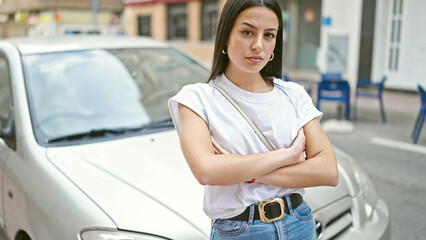 Wall Mural - Young beautiful hispanic woman standing with serious expression and arms crossed gesture by car at street