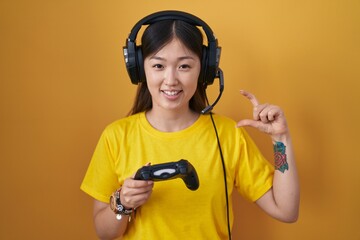 Poster - Chinese young woman playing video game holding controller smiling and confident gesturing with hand doing small size sign with fingers looking and the camera. measure concept.