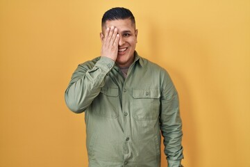 Wall Mural - Hispanic young man standing over yellow background covering one eye with hand, confident smile on face and surprise emotion.