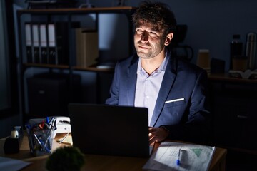 Poster - Hispanic young man working at the office at night smiling looking to the side and staring away thinking.