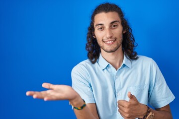 Poster - Young hispanic man standing over blue background showing palm hand and doing ok gesture with thumbs up, smiling happy and cheerful