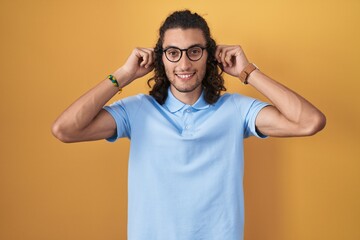 Poster - Young hispanic man standing over yellow background smiling pulling ears with fingers, funny gesture. audition problem