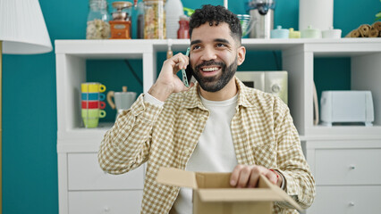 Canvas Print - Young hispanic man talking on smartphone unpacking cardboard box at dinning room