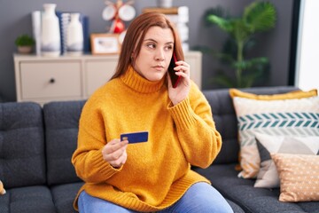 Wall Mural - Young beautiful plus size woman talking on smartphone holding credit card with serious expression at home