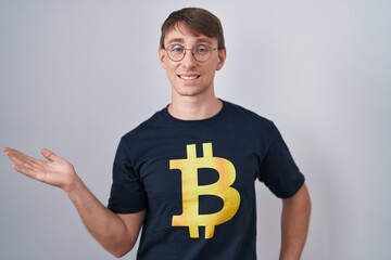 Sticker - Caucasian blond man wearing bitcoin t shirt smiling cheerful presenting and pointing with palm of hand looking at the camera.