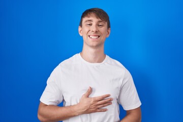 Poster - Caucasian blond man standing over blue background smiling and laughing hard out loud because funny crazy joke with hands on body.