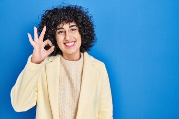 Poster - Young brunette woman with curly hair standing over blue background smiling positive doing ok sign with hand and fingers. successful expression.