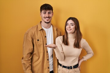 Wall Mural - Young hispanic couple standing over yellow background smiling cheerful presenting and pointing with palm of hand looking at the camera.