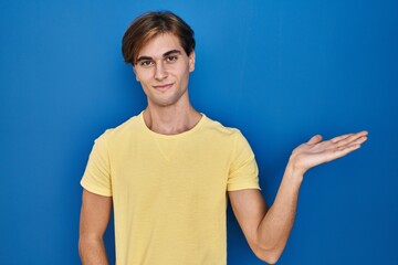 Wall Mural - Young man standing over blue background smiling cheerful presenting and pointing with palm of hand looking at the camera.