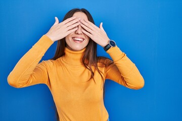 Canvas Print - Young brunette woman standing over blue background covering eyes with hands smiling cheerful and funny. blind concept.