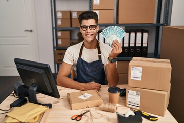 Sticker - Young hispanic man working at small business ecommerce holding brazilian reals looking positive and happy standing and smiling with a confident smile showing teeth